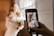 A person is holding a smartphone, capturing a photo of a bride dressed in a wedding gown. The bride stands near a window, holding a bouquet, with soft light illuminating her.