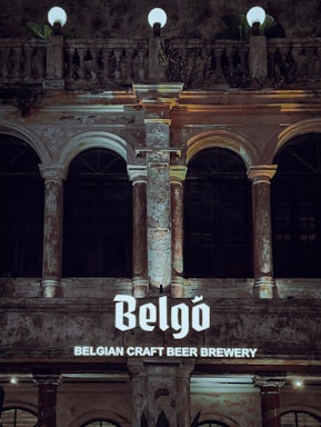 An ornate building facade featuring tall columns and arches, with textured, aged stonework and glowing spherical lamps on top. The facade is illuminated by a sign reading 'Belgo Belgian Craft Beer Brewery', displaying in a bold font.
