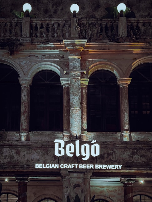 An ornate building facade featuring tall columns and arches, with textured, aged stonework and glowing spherical lamps on top. The facade is illuminated by a sign reading 'Belgo Belgian Craft Beer Brewery', displaying in a bold font.