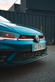 The image captures the front view of a blue Volkswagen GTI car, showcasing part of its grille and a visible license plate. The car's emblem and the GTI badge are prominent, with a backdrop of industrial containers adding a modern, urban setting.