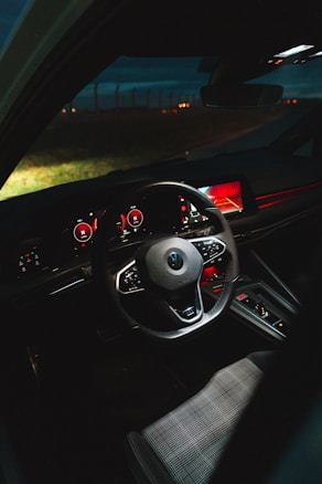 The interior of a car is illuminated dimly, highlighting a modern steering wheel, digital dashboard, and control panel with red accents. The environment outside the windshield appears to be a dark, open road with faint lights visible in the distance.