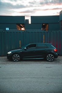 A sleek black car is parked on a gravel surface in front of a row of large, stacked shipping containers. The containers have a blue-gray color and are marked with yellow text. The sky is partially cloudy with a soft, warm light peeking through, suggesting either sunrise or sunset.