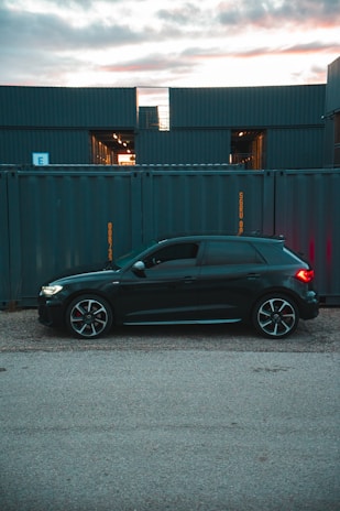 A sleek black car is parked on a gravel surface in front of a row of large, stacked shipping containers. The containers have a blue-gray color and are marked with yellow text. The sky is partially cloudy with a soft, warm light peeking through, suggesting either sunrise or sunset.