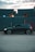 A sleek black car is parked on a gravel surface in front of a row of large, stacked shipping containers. The containers have a blue-gray color and are marked with yellow text. The sky is partially cloudy with a soft, warm light peeking through, suggesting either sunrise or sunset.