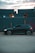 A sleek black car is parked on a gravel surface in front of a row of large, stacked shipping containers. The containers have a blue-gray color and are marked with yellow text. The sky is partially cloudy with a soft, warm light peeking through, suggesting either sunrise or sunset.