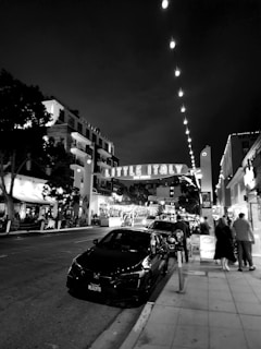 A nighttime street scene featuring a prominent sign reading 'Little Italy' in an urban area. The street is lined with cars, with people walking along the sidewalk. Buildings and streetlights are illuminated, creating a lively atmosphere.
