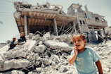 A young child is crying in the foreground, standing on rubble with a severely damaged building in the background. Two other children are visible climbing over the debris. The scene reflects chaos and destruction, possibly due to conflict or natural disaster.