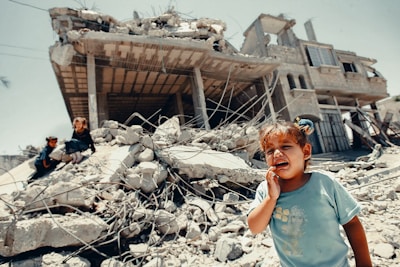 A young child is crying in the foreground, standing on rubble with a severely damaged building in the background. Two other children are visible climbing over the debris. The scene reflects chaos and destruction, possibly due to conflict or natural disaster.