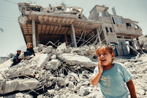 A young child is crying in the foreground, standing on rubble with a severely damaged building in the background. Two other children are visible climbing over the debris. The scene reflects chaos and destruction, possibly due to conflict or natural disaster.