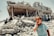 A young child is crying in the foreground, standing on rubble with a severely damaged building in the background. Two other children are visible climbing over the debris. The scene reflects chaos and destruction, possibly due to conflict or natural disaster.