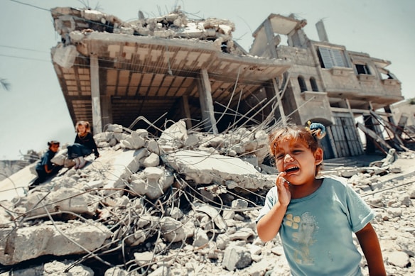 A young child is crying in the foreground, standing on rubble with a severely damaged building in the background. Two other children are visible climbing over the debris. The scene reflects chaos and destruction, possibly due to conflict or natural disaster.