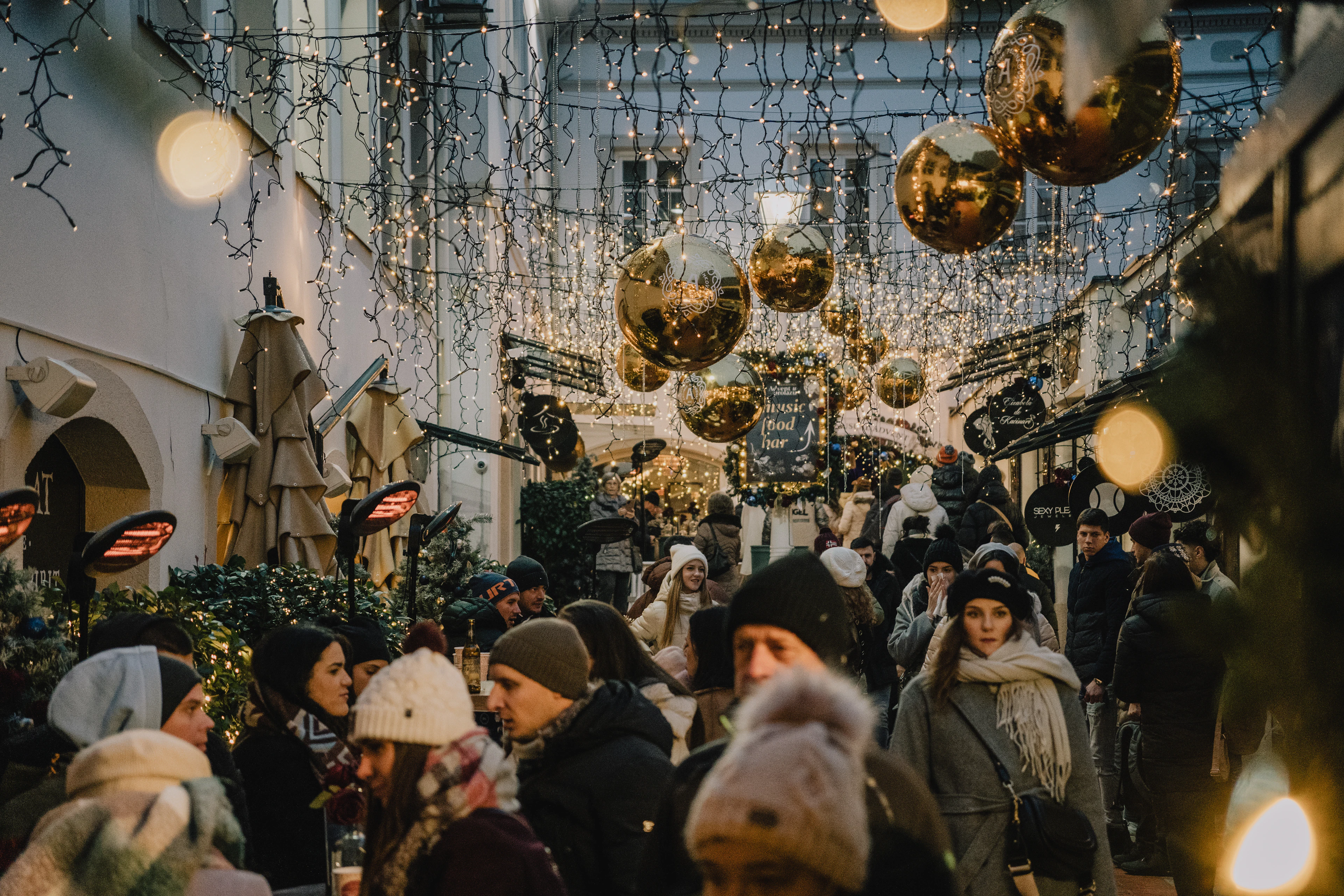 A covered passage strung with fairy lights and giant gold ornament baubles, crowded with visitors at a Christmas market.