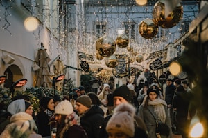 A busy street market adorned with sparkling lights and large golden ornaments, filled with people bundled in warm clothing. The atmosphere is festive and lively, with strings of fairy lights hanging overhead and shoppers milling about, engaging with vendors and enjoying the seasonal decorations.