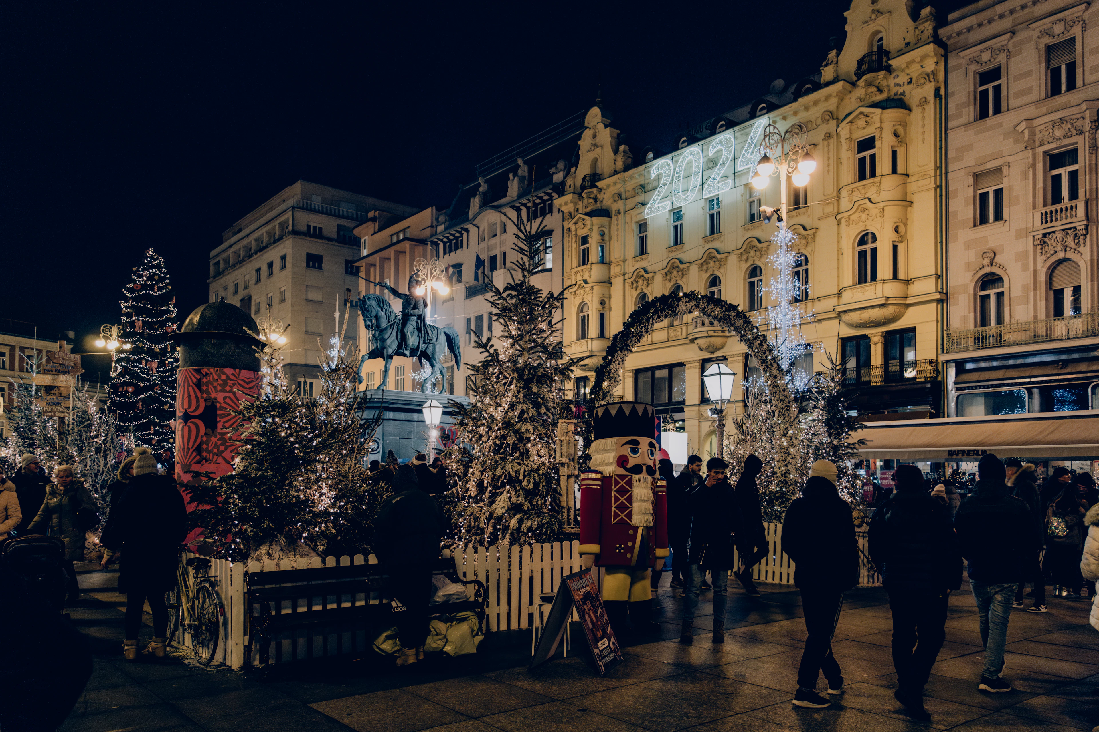 A festively lit square at night with Christmas trees, an illuminated arch, and a crowd gathered around a statue.