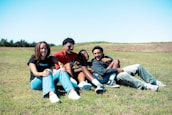 A group of four friends is sitting on the grass in an open field. They appear to be laughing and enjoying each other's company. The setting is outdoors under a clear blue sky. The grass is a bright, lively green, and the atmosphere seems relaxing and friendly.