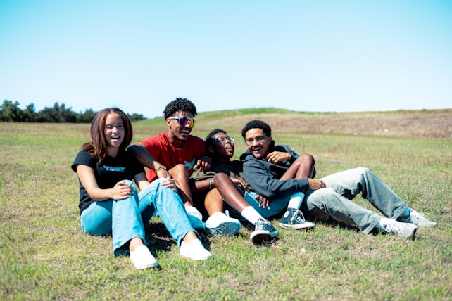 A group of four friends is sitting on the grass in an open field. They appear to be laughing and enjoying each other's company. The setting is outdoors under a clear blue sky. The grass is a bright, lively green, and the atmosphere seems relaxing and friendly.