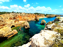 Brightly colored kayaks drift on clear, turquoise waters amidst dramatic, rocky cliffs with orange and beige hues. A vibrant, blue sky filled with fluffy white clouds forms the backdrop, adding to the scene's natural beauty.
