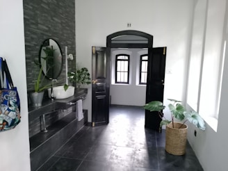 A modern bathroom features a dark tiled floor and wall with a round mirror above a sleek sink basin. Potted plants add a touch of greenery, and a colorful bag hangs on a hook. An open black door leads to a room with several windows letting in natural light.