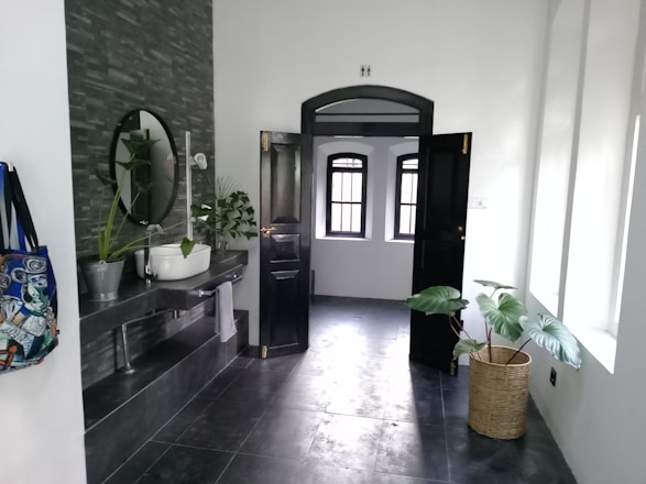 A modern bathroom features a dark tiled floor and wall with a round mirror above a sleek sink basin. Potted plants add a touch of greenery, and a colorful bag hangs on a hook. An open black door leads to a room with several windows letting in natural light.