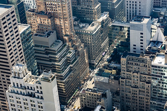 A dense urban landscape featuring a variety of high-rise buildings. The architecture includes a mix of modern and older styles, with glass facades and masonry. The streets below are bustling with traffic and people, and there is a visible network of streets and intersections.