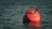A red buoy floating on the calm surface of the sea, tethered by a rope. The water is a deep greenish-blue and reflects light from the buoy.
