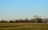 A vintage biplane flying low over an open grassy field with a backdrop of leafless trees under a clear blue sky. The aircraft has colorful markings on its body and tail.
