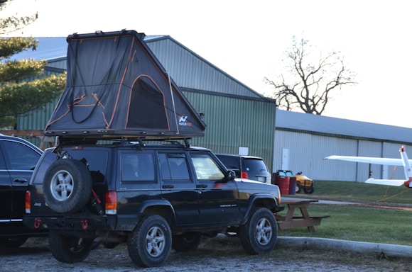 A dark-colored SUV with a tent mounted on its roof is parked in a lot. The tent is set up, and there are other vehicles nearby. In the background, there are hangar-like buildings and a small aircraft with its wing partially visible. The setting appears to be an outdoor area with some grass and sparse trees.