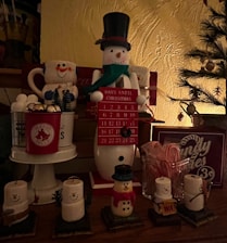 An arrangement of Christmas-themed decorations featuring a snowman countdown calendar in the center. The snowman is wearing a top hat and scarf, holding a red sign with the numbers to count down the days until Christmas. Surrounding it are various festive items, including mugs shaped like snowmen, miniature Christmas trees, candle holders resembling snowmen, and a jar filled with candy canes. The setting is warmly lit, enhancing the cozy holiday atmosphere.