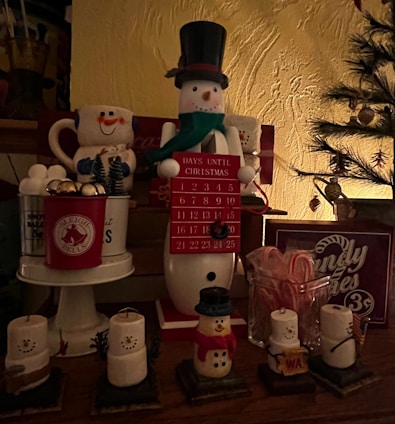 An arrangement of Christmas-themed decorations featuring a snowman countdown calendar in the center. The snowman is wearing a top hat and scarf, holding a red sign with the numbers to count down the days until Christmas. Surrounding it are various festive items, including mugs shaped like snowmen, miniature Christmas trees, candle holders resembling snowmen, and a jar filled with candy canes. The setting is warmly lit, enhancing the cozy holiday atmosphere.