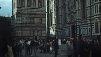 A large, ornate building with intricate details and patterns dominates the scene. The walls are adorned with geometric designs and tall, elegant windows. Many people are gathered around, some standing and others sitting, seemingly engrossed in observing the architectural features. The atmosphere is bustling with activity, as tourists explore the historical site.