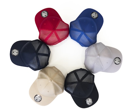 Six baseball caps are arranged in a circular pattern on a white background. The caps are of different colors including red, blue, gray, navy, beige, and black. Each cap features a round logo patch on the brim.