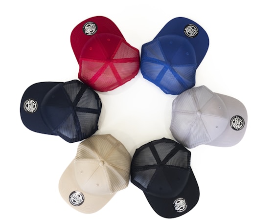 Six baseball caps are arranged in a circular pattern on a white background. The caps are of different colors including red, blue, gray, navy, beige, and black. Each cap features a round logo patch on the brim.