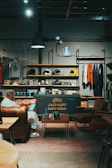 A cozy interior of a cafe features a mix of industrial and vintage design, with leather seating, a table with a drink, and a person using a laptop. The shelves are stocked with merchandise such as helmets and t-shirts. The lighting is warm and ambient, contributing to the inviting atmosphere.