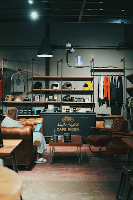 A cozy interior of a cafe features a mix of industrial and vintage design, with leather seating, a table with a drink, and a person using a laptop. The shelves are stocked with merchandise such as helmets and t-shirts. The lighting is warm and ambient, contributing to the inviting atmosphere.