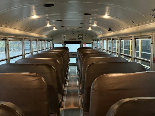The image depicts the interior of an empty school bus, characterized by rows of brown seats and large windows on either side. The ceiling is curved with overhead lighting, and there are no passengers or objects visible on the seats.
