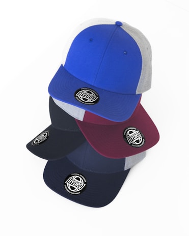 Multiple baseball caps are stacked in an overlapping manner. The caps feature a round sticker with the words 'HYDRO' and 'DESIGNED IN AUSTRALIA'. The top cap is blue with gray panels, while the others in the stack include different shades like maroon and black.