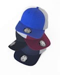 Multiple baseball caps are stacked in an overlapping manner. The caps feature a round sticker with the words 'HYDRO' and 'DESIGNED IN AUSTRALIA'. The top cap is blue with gray panels, while the others in the stack include different shades like maroon and black.