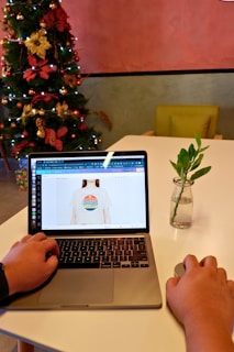 A person is using a laptop on a white table with a minimalistic setup. The laptop screen displays an image of a t-shirt with colorful text on it. To the right, there's a small vase containing a plant with a few green leaves. In the background, there's a decorated Christmas tree with red and gold ornaments and lights. The wall behind the setup is painted in muted pink and green tones, and there is a green chair visible.