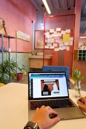 A hand is operating a laptop on a white table in a creatively styled room. The room features pink and green walls with a corkboard filled with various papers and notes. Potted plants and minimal decor give the space a casual and modern feel. Overhead, there are hanging lights, and a vase with a small plant is visible on the table.