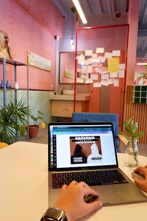 A hand is operating a laptop on a white table in a creatively styled room. The room features pink and green walls with a corkboard filled with various papers and notes. Potted plants and minimal decor give the space a casual and modern feel. Overhead, there are hanging lights, and a vase with a small plant is visible on the table.