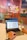 Hands typing on a laptop in a colorful and creative office space. The laptop screen displays an image of a T-shirt with 'Babe' written on it. The desk also has a small plant in a glass bottle. The background shows a bulletin board with various notes and a mirror above a sink.