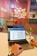 Hands typing on a laptop in a colorful and creative office space. The laptop screen displays an image of a T-shirt with 'Babe' written on it. The desk also has a small plant in a glass bottle. The background shows a bulletin board with various notes and a mirror above a sink.