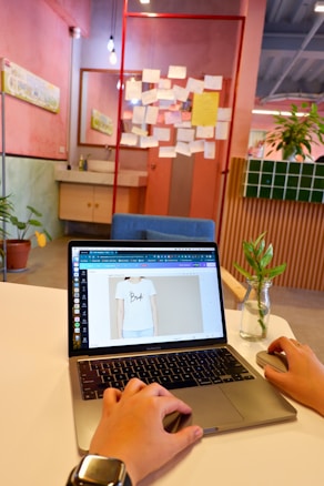 Hands typing on a laptop in a colorful and creative office space. The laptop screen displays an image of a T-shirt with 'Babe' written on it. The desk also has a small plant in a glass bottle. The background shows a bulletin board with various notes and a mirror above a sink.