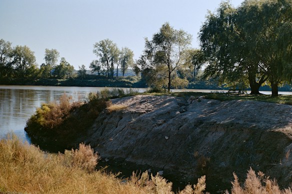 A serene riverbank scene with calm water reflecting the light. Trees with lush green leaves line the opposite bank, and a grassy area with a picnic table is shaded by a large tree. The foreground features a rocky, slightly eroded area leading down to the water.