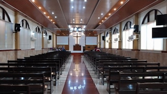The interior of a church with rows of wooden pews leading up to a pulpit. A large cross is prominently displayed on the front wall above the pulpit. The ceiling is adorned with recessed lighting, and there are arched windows along the sides. The floor is polished, reflecting the lights from above.