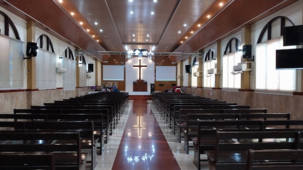 The interior of a church with rows of wooden pews leading up to a pulpit. A large cross is prominently displayed on the front wall above the pulpit. The ceiling is adorned with recessed lighting, and there are arched windows along the sides. The floor is polished, reflecting the lights from above.