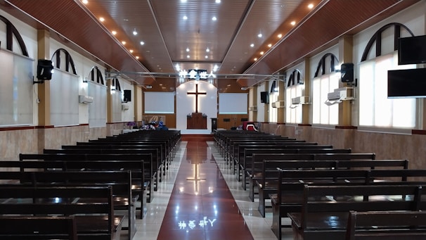 The interior of a church with rows of wooden pews leading up to a pulpit. A large cross is prominently displayed on the front wall above the pulpit. The ceiling is adorned with recessed lighting, and there are arched windows along the sides. The floor is polished, reflecting the lights from above.