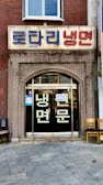 A storefront with a brick facade and a large sign in Korean characters above the entrance. There are glass doors with more Korean writing and a couple of chairs placed outside on the tiled pavement.