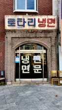 A storefront with a brick facade and a large sign in Korean characters above the entrance. There are glass doors with more Korean writing and a couple of chairs placed outside on the tiled pavement.