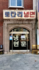 A storefront with a brick facade and a large sign in Korean characters above the entrance. There are glass doors with more Korean writing and a couple of chairs placed outside on the tiled pavement.